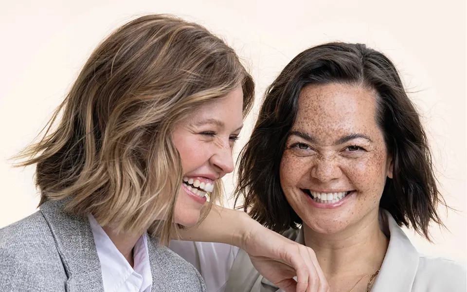 Zwei Frauen mit kurzen Haaren lächeln im Nahporträt, eine trägt grauen Blazer, die andere hat Sommersprossen, heller Hintergrund.